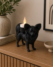 Zwarte Papillon hond waxinelichthouder met brandend waxinelicht op houten meubel in warm interieur.