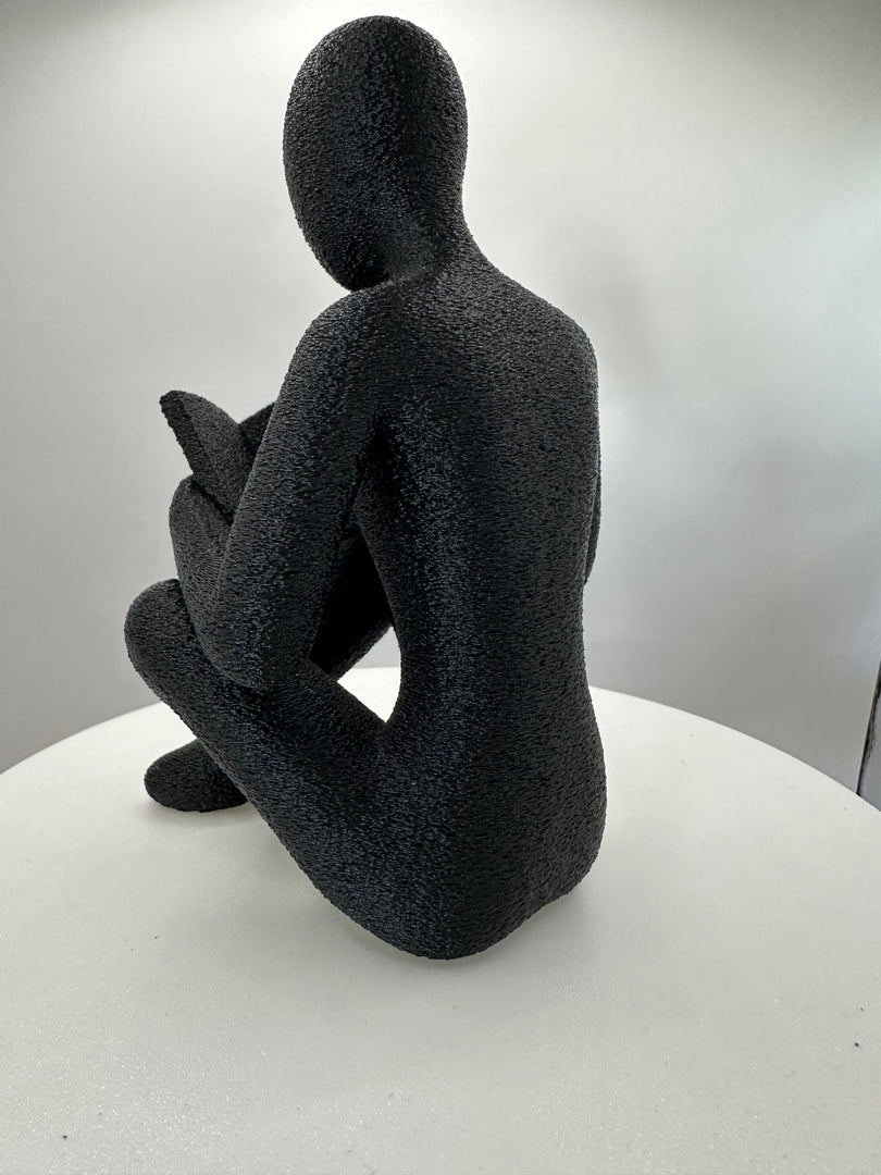 Minimalistische sculptuur van een lezende figuur met boek gezien vanuit zijprofiel.