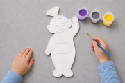 Kind schildert een verfbaar Tinky Winky figuur uit de Teletubbies met meegeleverde verf in paars, geel en grijs en een penseel van Hunoba Creative.
