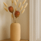 Beige Siervaas Rosella Chic met proteabloemen en bunny tails op een houten kastje bij een deurstijl.