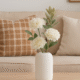 Witte Siervaas Rosella Chic met witte kunstbloemen en blaadjes op een houten tafel voor een beige bank.