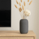 Grijze Siervaas Rosella Chic met droogbloemen op een houten tv-meubel naast een flatscreen.