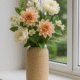 Beige Siervaas Rosella Chic met perzik- en crèmekleurige kunstbloemen op een vensterbank.